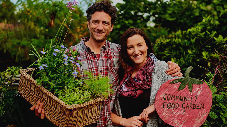 Two smiling growers holding a basket of vegetables beside a Community Food Garden sign
